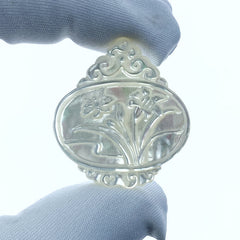 A hand-carved white mother of pearl pendant featuring a floral design with orchids and butterflies, held in a gloved hand.