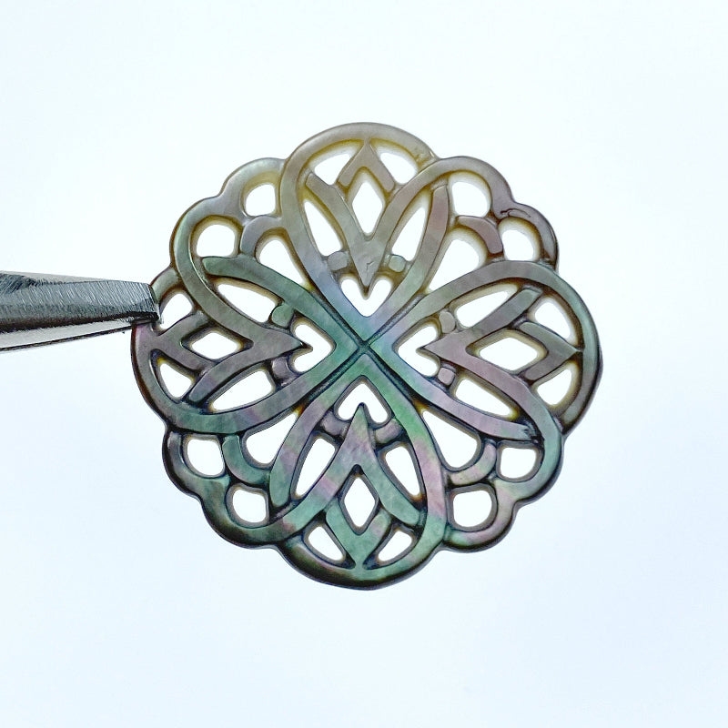 A close-up image of a round black mother of pearl filigree pendant with a metallic sheen, held by a needle.