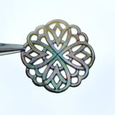 A close-up image of a round black mother of pearl filigree pendant with a metallic sheen, held by a needle.