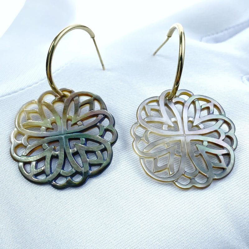 A close-up image of a round black mother of pearl filigree pendant with a metallic sheen, held by a needle.