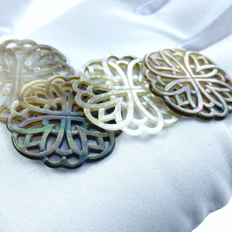 A close-up image of a round black mother of pearl filigree pendant with a metallic sheen, held by a needle.