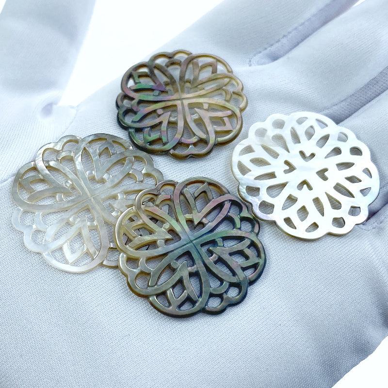 A close-up image of a round black mother of pearl filigree pendant with a metallic sheen, held by a needle.