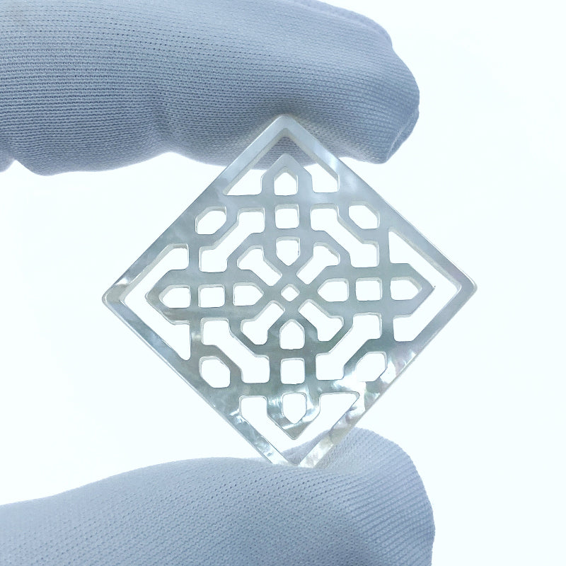 A hand-carved white mother of pearl filigree square pendant held between fingers, with a visible organic pattern.